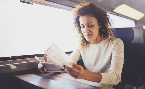 Enjoying business travel concept. Young beautiful brunette tourist girl travelling on the train sitting near the window using smartphone,holding ticket hands. - Foto, imagen