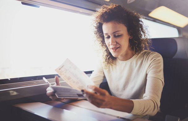 Enjoying business travel concept. Young beautiful brunette tourist girl travelling on the train sitting near the window using smartphone,holding ticket hands. - Foto, Imagen
