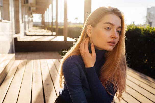 beautiful blond model girl in blue sweater sits outdoors in the sun, posing - Photo, Image