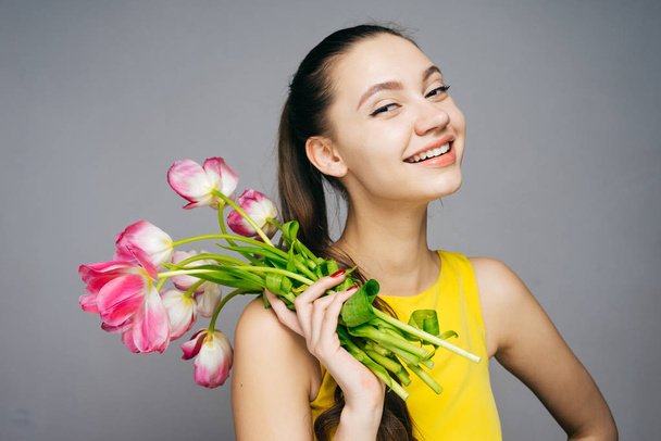 happy young girl in yellow dress holds pink flowers, laughs and looks at camera - Photo, Image