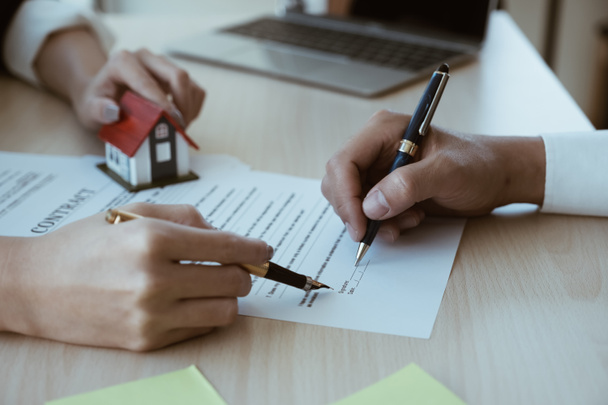 Beschnittenes Bild eines Immobilienmaklers, der dem Kunden hilft, am Schreibtisch mit Hausmodell ein Vertragspapier zu unterzeichnen - Foto, Bild