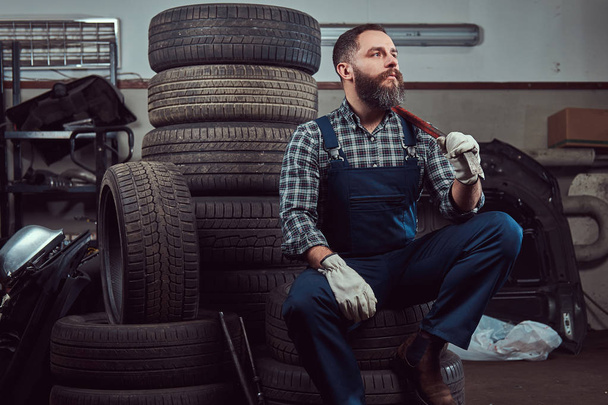 bärtiger Mechaniker in Uniform, hält ein Brecheisen in der Hand, sitzt auf alten Autoreifen in der Garage. - Foto, Bild