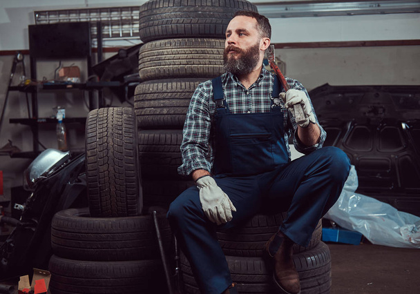 Brodaty mechanik, ubrany w mundur, kryje w sobie Łom, siedzi na zużytych opon samochodowych w garażu. - Zdjęcie, obraz