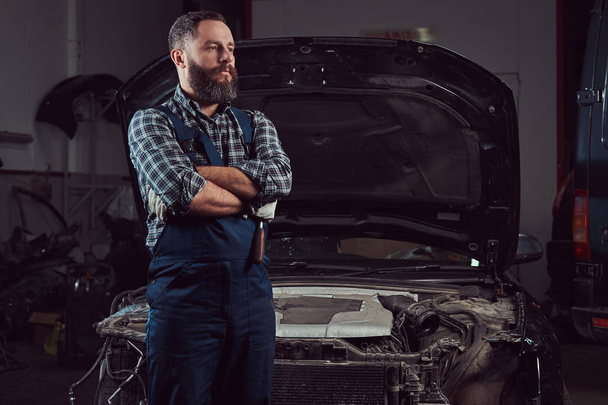 Бородатий експерт-механік, одягнений в уніформу, стоїть з схрещеними руками проти автомобіля в гаражі
. - Фото, зображення