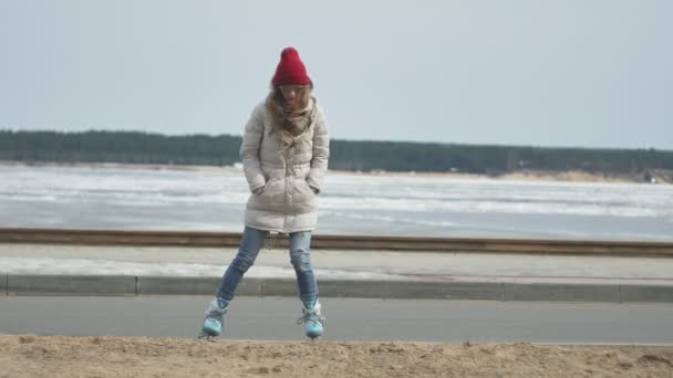 Fiatal gyönyörű nő egy piros sapkát visel, sportos meleg ruhát és a görgők, lovaglás a tengerparton úton - Felvétel, videó