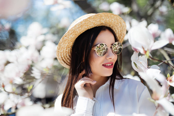 Young woman in sunglasses and straw hat surrounded with magnolia. Spring season - Foto, Imagen