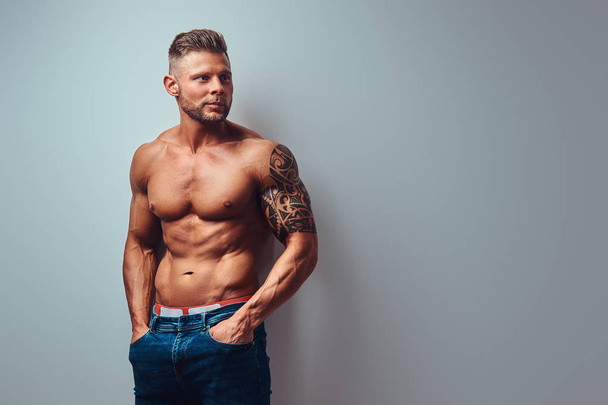A handsome shirtless bodybuilder with stylish haircut and beard, with tattoo on his arm, posing in a studio. Isolated on a gray background. - Foto, imagen