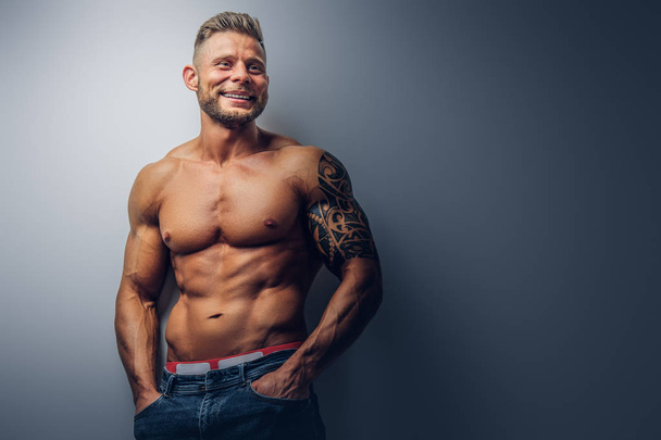 Smiling handsome shirtless bodybuilder with stylish haircut and beard, with tattoo on his arm, posing in a studio. Isolated on a gray background. - Foto, Bild