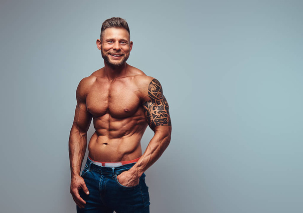 Smiling handsome shirtless bodybuilder with stylish haircut and beard, with tattoo on his arm, posing in a studio. Isolated on a gray background. - Photo, Image