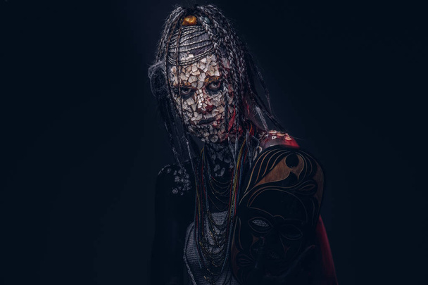 Πορτρέτο του ένα τρομακτικό αφρικανικός σαμάνος θηλυκό με ένα απολιθωμένο ραγισμένα δέρμα και dreadlocks σε σκούρο φόντο. Έννοια του μακιγιάζ. - Φωτογραφία, εικόνα