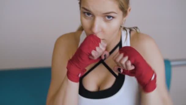 Die junge Frau boxt, die Hände in rote Boxbänder gehüllt. Schauen Sie in die Kamera. Schöne Boxerin beim Training im Fitnessstudio. - Filmmaterial, Video