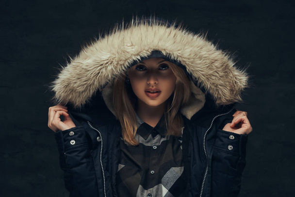 Portrait of a sensual blonde girl in a winter jacket and fur hood, standing in a studio. Isolated on a dark background. - Foto, Imagem