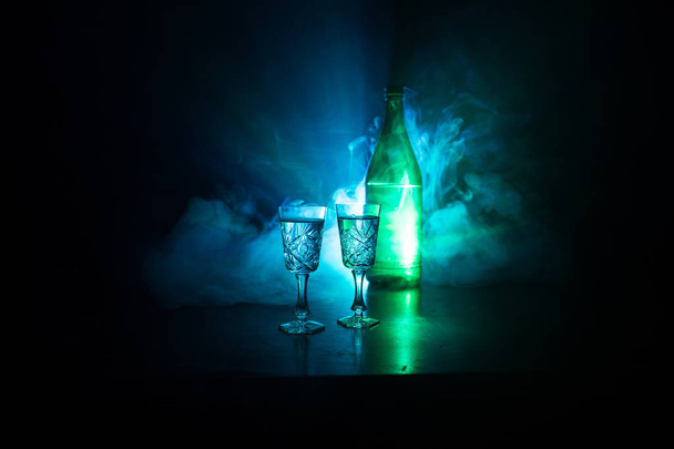 zwei Gläser Wodka mit Flasche auf dunklem, nebeligen Hintergrund mit leuchtenden Lichtern (Laser, Herd) mehrfarbig. Club Drinks Thema Dekoration. Leere Räume. Selektiver Fokus - Foto, Bild