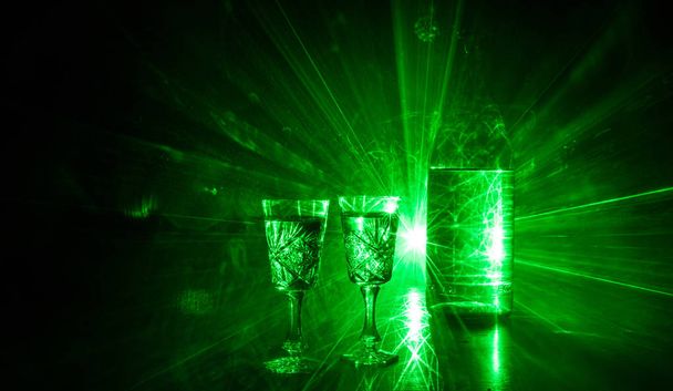 Δύο ποτήρια βότκα με μπουκάλι σε σκοτεινό, ομιχλώδη club στυλ φόντου με λαμπερό παραδεισένιους φώτα (Laser, Stobe). Λέσχη ποτά θέμα διακόσμησης. Κενό χώρο. Επιλεκτική εστίαση - Φωτογραφία, εικόνα
