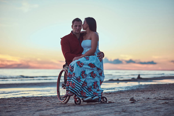 A loving couple, a woman sits on her husbands lap, resting on a beach against a background of a bright dawn. - Photo, Image