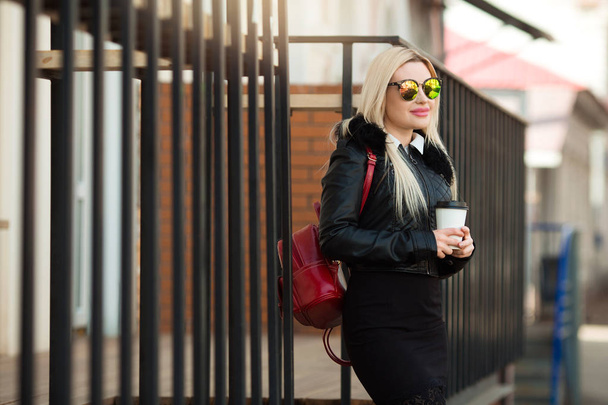 beautiful young girl in a black jacket and glasses on the street with a cup of coffee in hands - 写真・画像