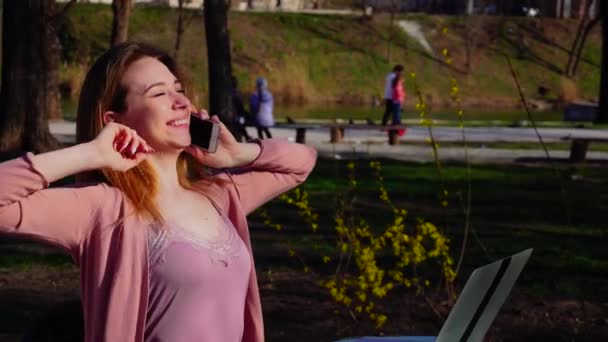 Gelukkige vrouwelijke persoon spreken door smartphone in park en werken met laptop. - Video