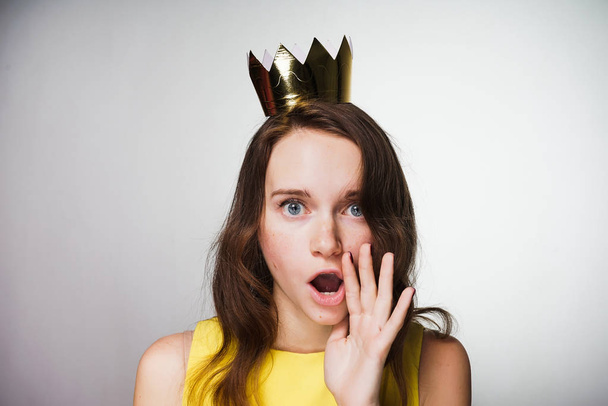 A young blue-eyed girl in a yellow dress looks surprised, a golden crown on her head - Photo, Image