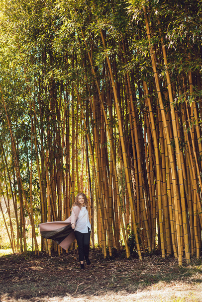 stylish young woman in pink coat walking near tall bamboo trees in park - Photo, image