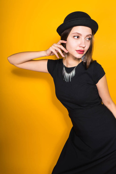 confident attractive girl in a long black dress and hat posing on a yellow background, in expensive earrings - Fotoğraf, Görsel
