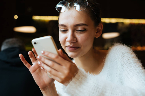 beautiful young girl student in a white jacket and glasses sits in a cafe and looks into her smartphone - Фото, изображение