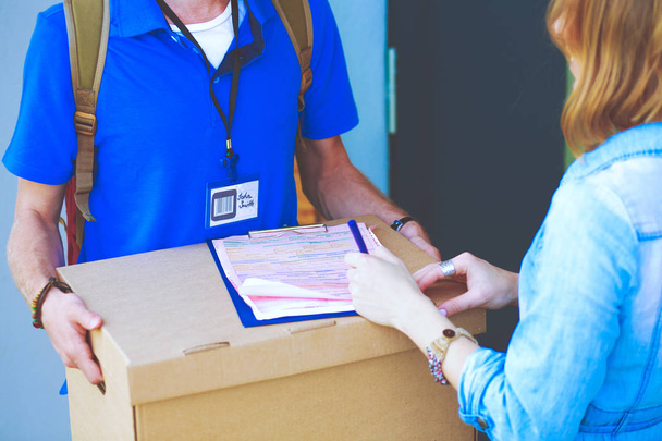 Hymyilevä postimies sinisessä univormussa toimittamassa pakettilaatikkoa vastaanottajalle kuriiripalvelun konsepti. Hymyilevä lähetti sinisessä univormussa. - Valokuva, kuva