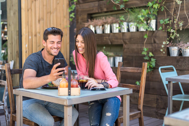 Happy young couple seating in a restaurant terrace with a smartphone - Фото, изображение