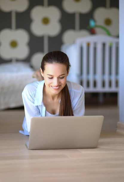 Young woman sitting on the floor near childrens cot with laptop. Young mom - Zdjęcie, obraz
