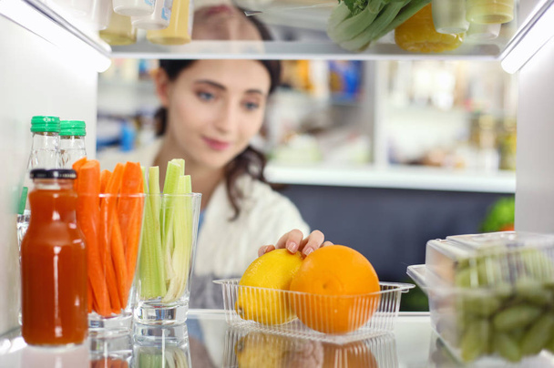 健康的な食べ物、野菜や果物でいっぱいのオープン冷蔵庫の近くに立っている女性の肖像画。女性像 - 写真・画像