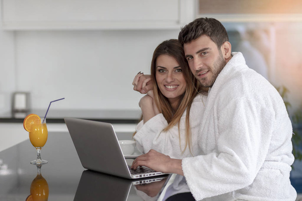 young couple in bathrobe at home having coffee in the kitchen and working on laptop computer - Photo, image
