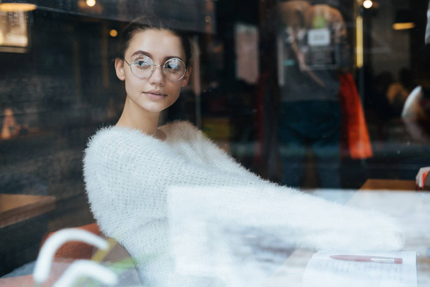 young beautiful girl student in glasses sits in a cafe after studying at university and looks out the window - Valokuva, kuva