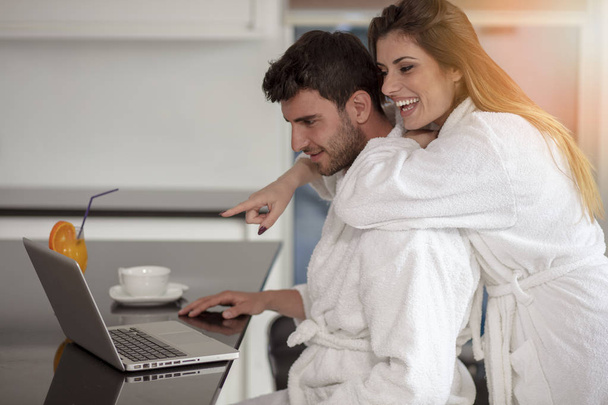 young couple in bathrobe at home having coffee in the kitchen and working on laptop computer - Photo, image