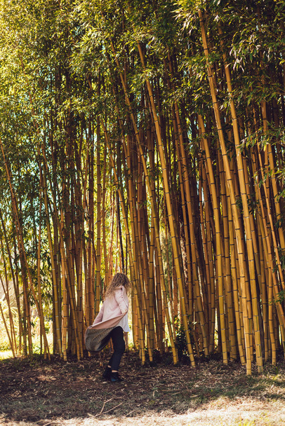 curly stylish woman in pink coat posing next to tall bamboo trees, spring weather - Foto, immagini