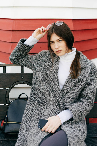 dark haired city girl in a trendy gray coat sitting on a bench outdoors, street style - Фото, изображение