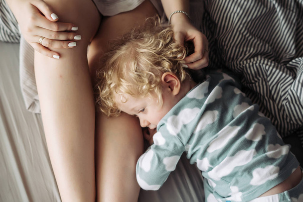 curly little baby boy hugs mom by the leg, lie on the bed and rest - Φωτογραφία, εικόνα