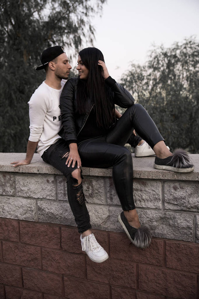 casal apaixonado: uma bela jovem em roupas pretas sorrindo abraçando um jovem elegante sentado em um parapeito de concreto em um parque em um fundo de edifícios altos à distância
 - Foto, Imagem