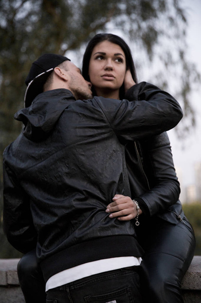 Couple in love: a beautiful young girl in a black leather jacket and pants gently hugs a stylish young man sitting on a concrete parapet in a park against a backdrop of tall buildings in the distance - Foto, afbeelding