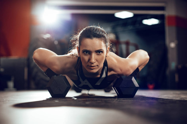 Mujer muscular joven haciendo ejercicio push-up con dumbell en el entrenamiento duro en el gimnasio
 - Foto, Imagen
