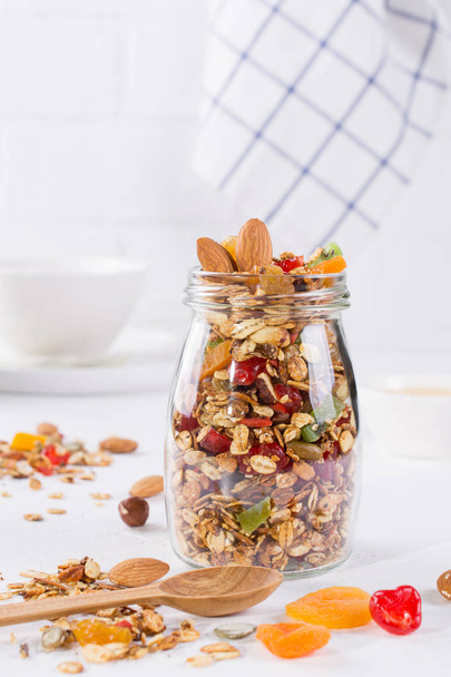 Glass jar with baked granola on white background. Oat flakes, honey, nuts, dried fruit and seeds. Healthy snak - Photo, Image