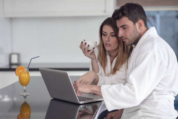 young couple in bathrobe at home having coffee in the kitchen and working on laptop computer - Foto, imagen