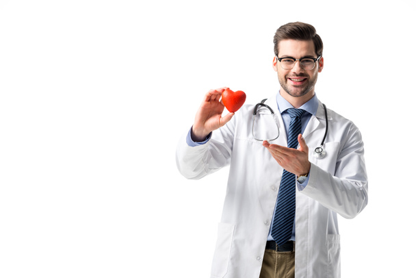Lachende arts dragen witte vacht met de stethoscoop en houden speelgoed hart geïsoleerd op wit - Foto, afbeelding