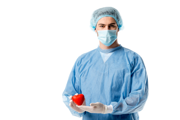 Cirujano en uniforme médico azul y máscara médica y celebración de corazón de juguete aislado en blanco
 - Foto, Imagen