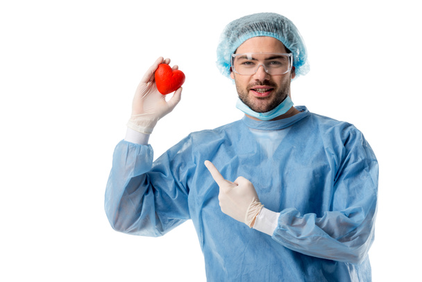 Cirujano sonriente en uniforme médico azul y máscara médica y apuntando al corazón del juguete aislado en blanco
 - Foto, Imagen