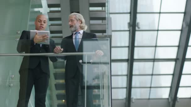 two corporate executives standing on second floor of a glass and steel building discussing business using digital tablet. - Footage, Video