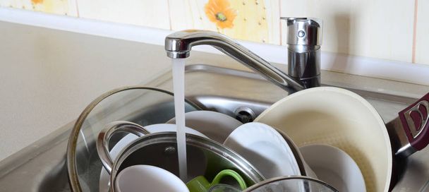 Piszkos edények és mosatlan konyhai eszközök hevernek habos vízben a csapvíz alatt egy konyhai csapból. - Fotó, kép
