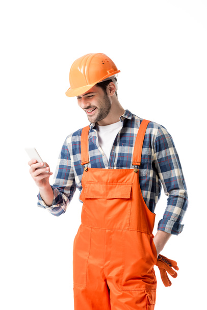 Smiling handyman in orange overall and helmet using smartphone isolated on white - Zdjęcie, obraz