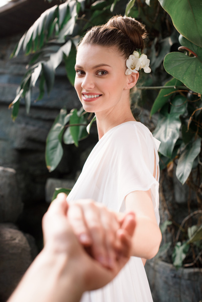 χαμογελαστή γυναίκα στην Λευκή φόρεμα με λουλούδι στα μαλλιά, κρατώντας τα χέρια με τον άνθρωπο - Φωτογραφία, εικόνα
