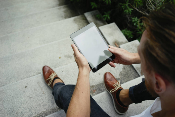 schöner junger Mann benutzt digitales Tablet während er auf der Treppe sitzt - Foto, Bild