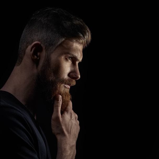 Atraente jovem pensativo olha para a distância acariciando sua barba
 - Foto, Imagem