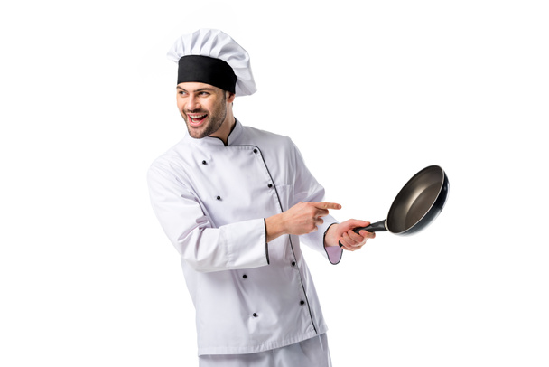 smiling chef in uniform pointing at frying pan in hand isolated on white - Fotó, kép